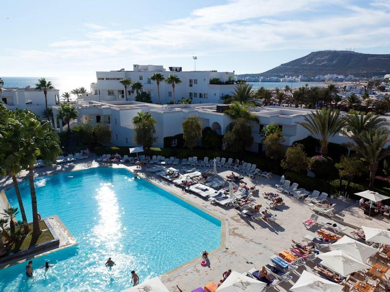 Großer Hotelpool mit vielen Sonnenliegen, Palmen und weißer Hotelanlage vor Hügelkulisse.