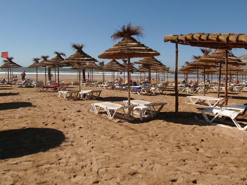 Strand mit Sonnenliegen und Strohdächern unter blauem Himmel, sandiger Boden, sonnig.