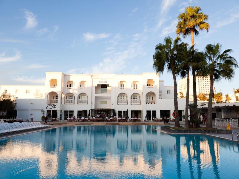 Weißes Hotel mit Balkonfenstern und großem Pool, umgeben von hohen Palmen bei blauem Himmel.