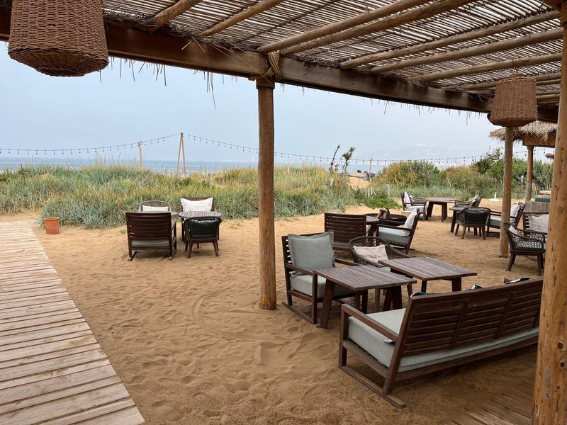 Strand mit Holzsteg, Sandboden, Sitzgruppen aus Holzmöbeln und überdachtem Bereich aus Bambus.