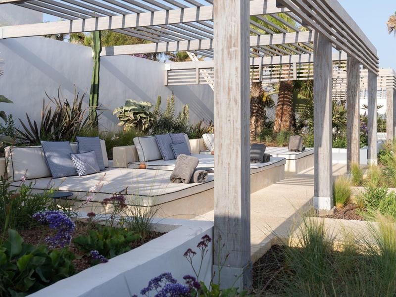 Moderner Außenbereich mit grauem Pergola, weißem Sofa, grünen Pflanzen und blühenden Blumen.