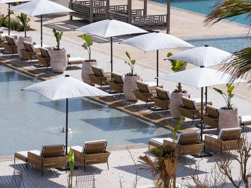 Strand mit blauen Liegestühlen und weißen Sonnenschirmen am Wasser mit Palmen im Hintergrund.