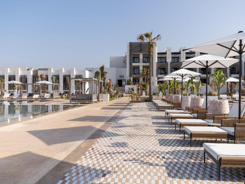 Moderner Hotelpool mit Liegestühlen und Sonnenschirmen neben einem weißen Gebäude bei klarem Himmel.