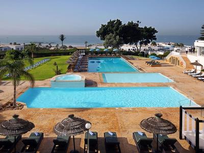 Hotelpool mit klar blauem Wasser, Liegestühlen und Blick auf das Meer im Hintergrund.