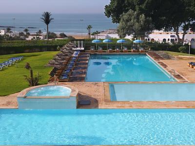Hotelpoolbereich mit mehreren Pools, Liegestühlen und Blick auf das Meer im Hintergrund.