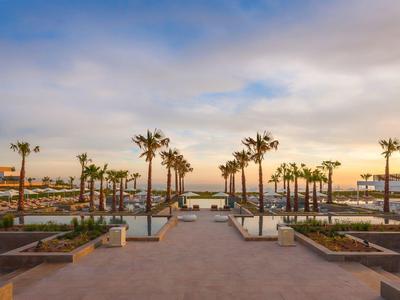 Weite Promenade mit Palmen, Treppen und Sonnenuntergang über einem Hafen mit Booten.