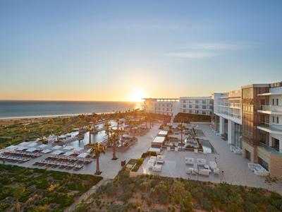 Západ slunce nad moderním hotelovým komplexem s venkovním bazénem a výhledem na moře.