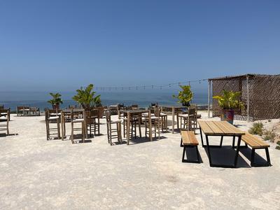 Venkovní posezení plážové restaurace s dřevěným nábytkem a výhledem na moře pod jasnou oblohou.