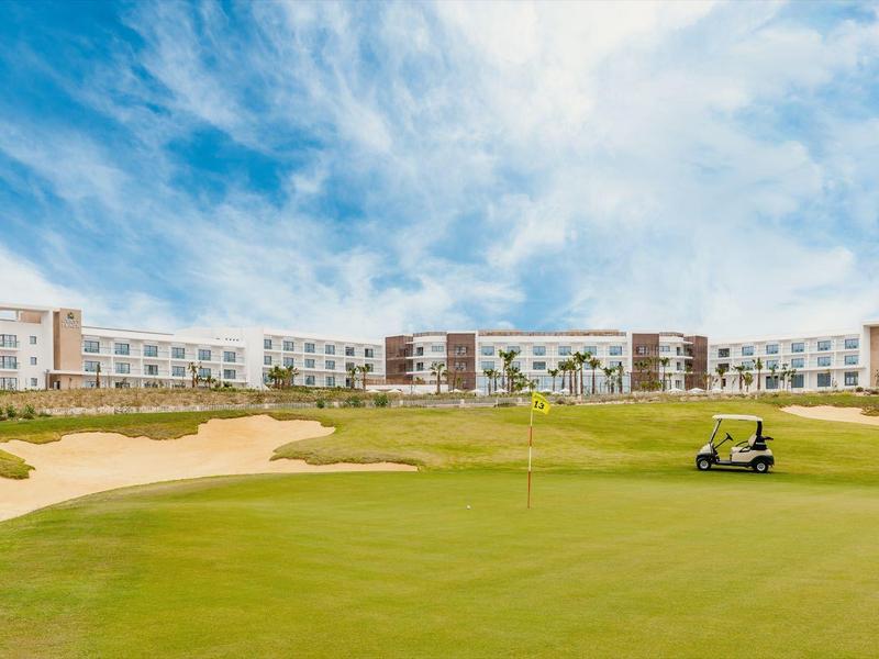 Großer Golfplatz mit weitläufigem Grün, Bunkern und einem Gebäude im Hintergrund unter blauem Himmel.
