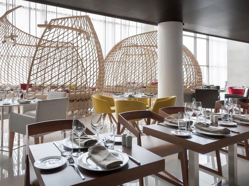 Modernes Restaurant mit Holztischen, weißen Stühlen und geflochtenen Hängesesseln neben großen Fenstern.