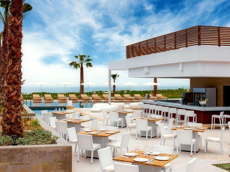 Strandrestaurant mit weißen Stühlen, Tischen, Palmen und Blick auf das blaue Meer.