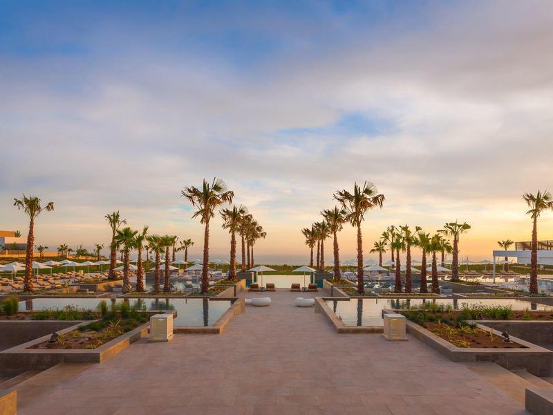 Weite Promenade mit Palmen, Treppen und Sonnenuntergang über einem Hafen mit Booten.
