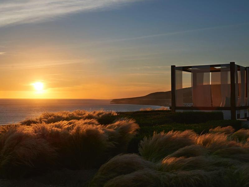 Sonnenuntergang über dem Meer, mit Gräsern im Vordergrund und einer offenen Holzkonstruktion rechts.