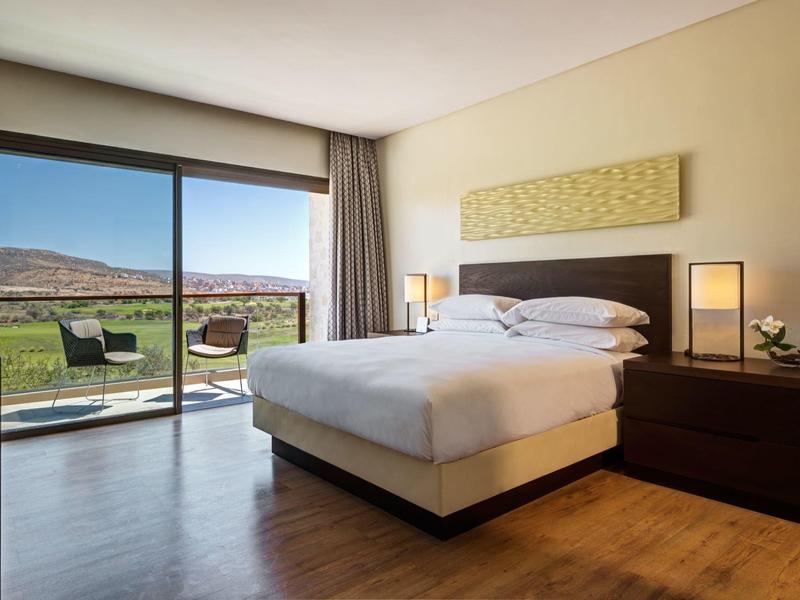 Modernes Schlafzimmer mit Doppelbett, großen Fenstern und Blick auf eine Terrasse und karge Landschaft.