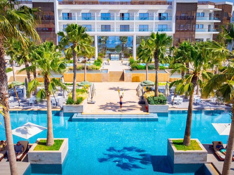 Moderner Poolbereich mit Palmen und Liegestühlen vor einem weißen Hotelgebäude bei Sonnenschein.