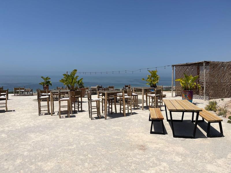 Leerer Außenbereich mit Holztischen und Bänken auf hellem Sand vor blauem Himmel und Meer.