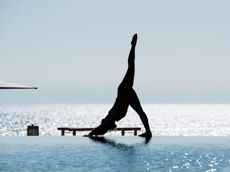 Person macht Yoga-Pose auf Poolrand, blauer Himmel, glänzendes Wasser im Hintergrund.