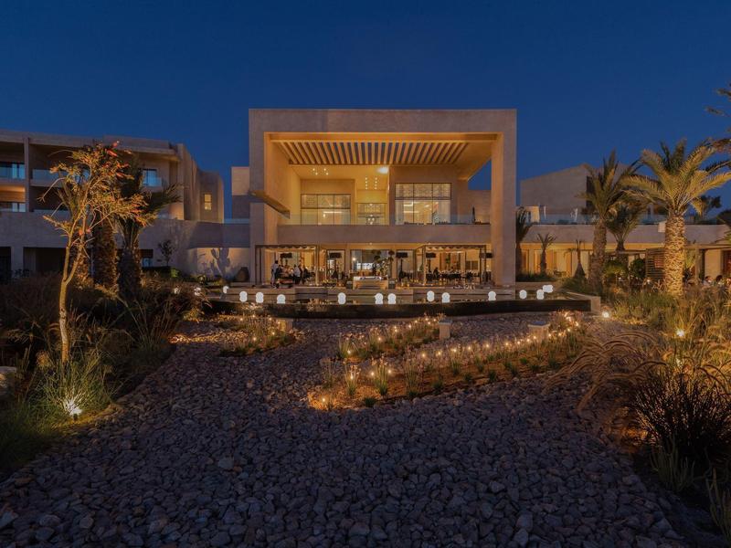 Modernes beleuchtetes Gebäude mit Holzstruktur und Pflanzen im Vordergrund bei Nacht.