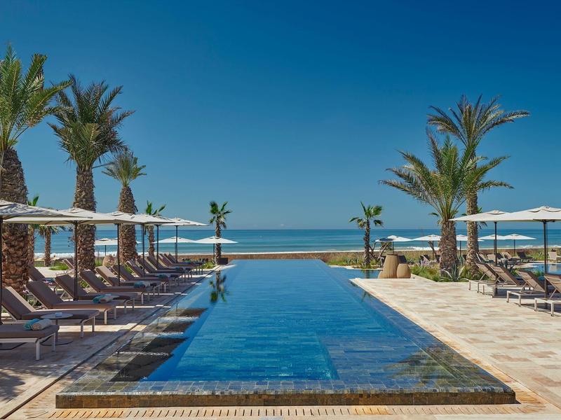 Infinity-Pool mit Sonnenliegen und Palmen, klarer Himmel, Blick auf ruhiges Meer am Horizont.
