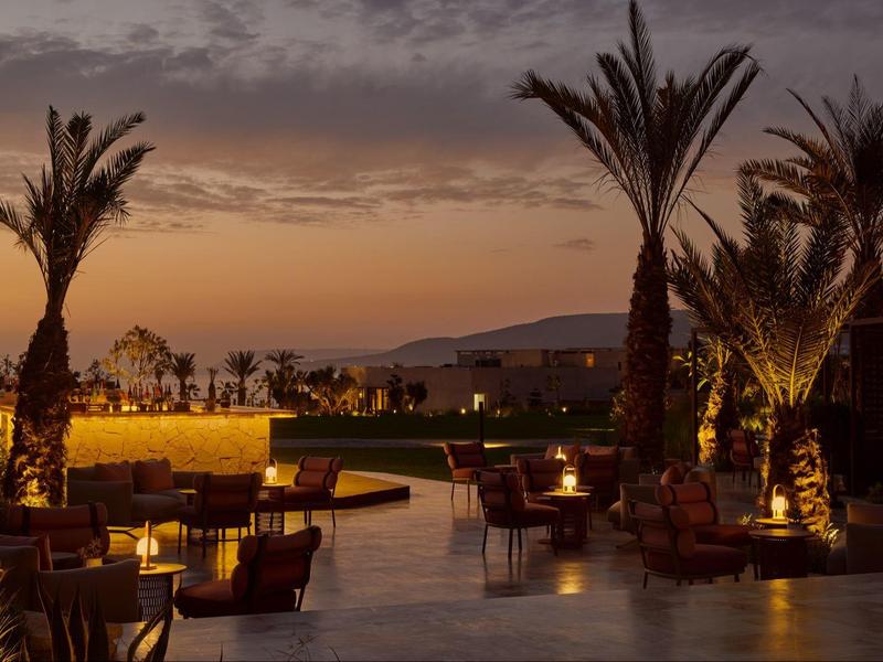 Abendliche Terrasse mit Palmen und warmem Licht vor Bergen und bewölktem Himmel.