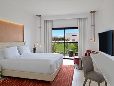 Nowoczesny pokój hotelowy z dużym łóżkiem, częścią wypoczynkową, czerwonym dywanem i widokiem na zielony ogród.