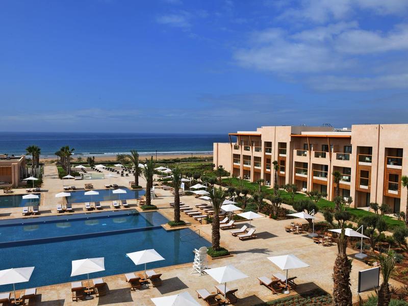 Modern hotel complex with pool, umbrellas, and palm trees by the sea under a blue sky.
