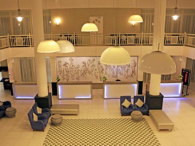 Heller, moderner Hotel-Lounge-Bereich mit Sitzgruppen, Teppich und großen Hängelampen.