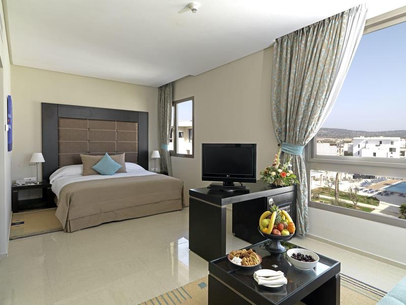 Nowoczesny pokój hotelowy z łóżkiem podwójnym, telewizorem, stolikami nocnymi i dużym oknem z widokiem na miasto.