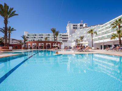 Large hotel pool with palm trees and lounge chairs under clear blue sky.