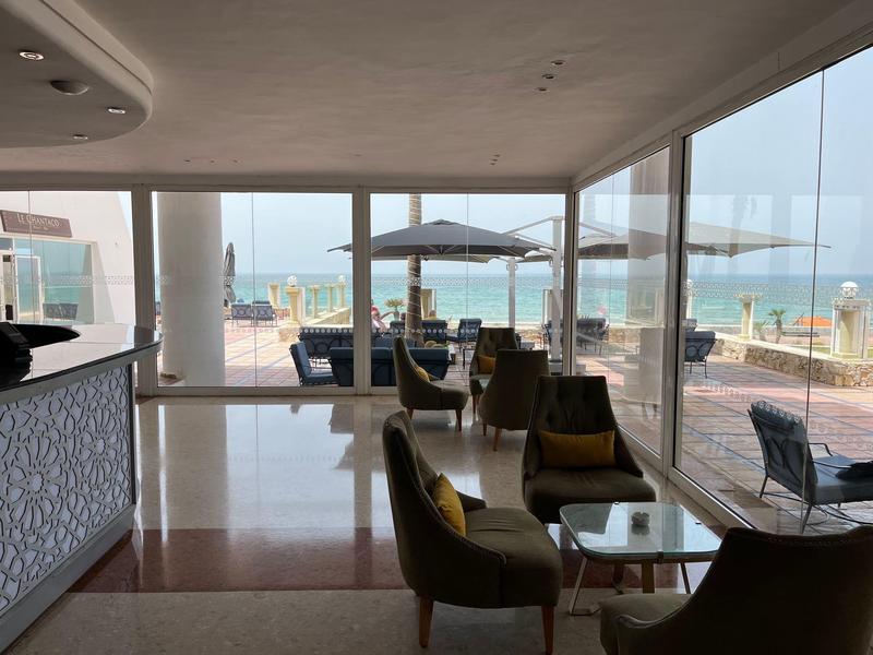 Hotel lounge met fauteuils en glazen tafels, uitzicht op terras en zee door grote ramen.