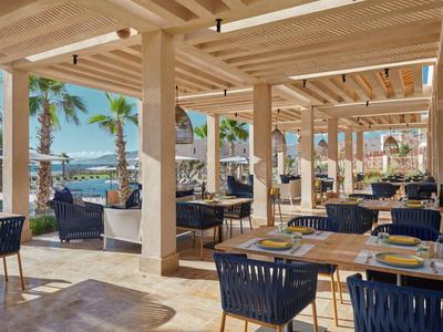 Offenes Restaurant mit Holzdach und Blick auf Palmen und Meer bei Sonnenschein.