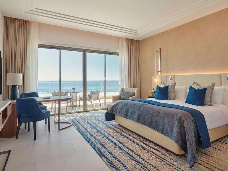 Lichte hotelkamer met groot bed, balkon en zeezicht in zonlicht.