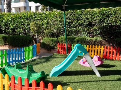 Kinderspielplatz mit bunter Umzäunung, Rutschen und grünem Kunstrasen vor Hotelgebäuden.