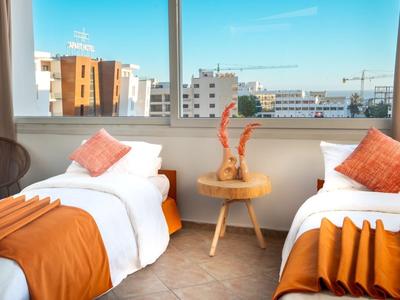 Hell eingerichtetes Hotelzimmer mit zwei Betten, orangenen Decken und Kissen nahe eines großen Fensters.