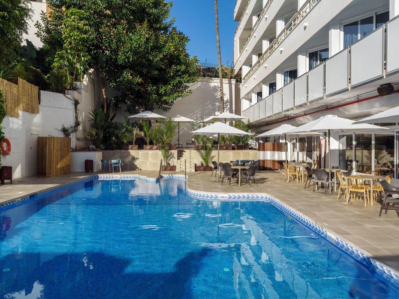 Ein blauer Pool mit weißen Stühlen und Sonnenschirmen neben einem mehrstöckigen Gebäude.