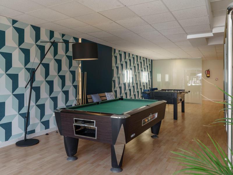 Innenraum mit Billardtisch, Tischfußballspiel und gemusterten Wänden, heller Holzboden, Deckenlampen.