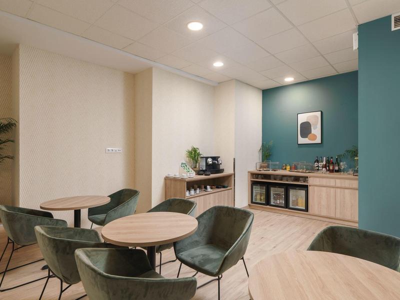 Moderner Besprechungsraum mit runden Holztischen, grünen Stühlen und Kaffeeecke vor türkisfarbener Wand.