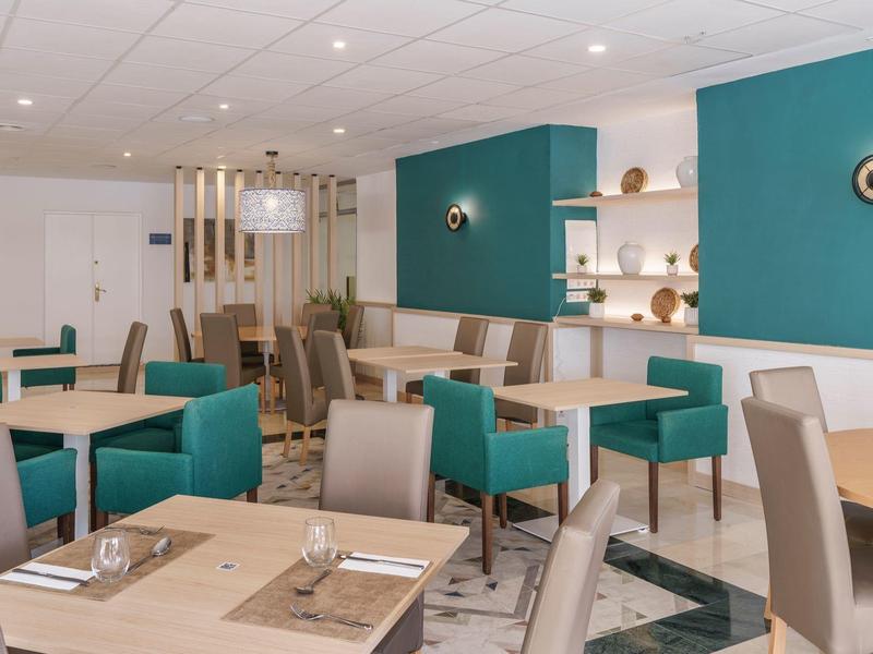 Moderner Speisesaal mit türkisfarbenen Wänden, hellen Tischen und grauen Sesseln, sauber und ordentlich.