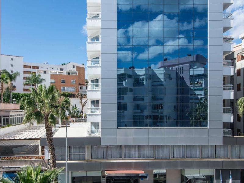 Modernes Glasgebäude mit Wolkenspiegelung, umgeben von Autos und Palmen, unter klarem blauem Himmel.