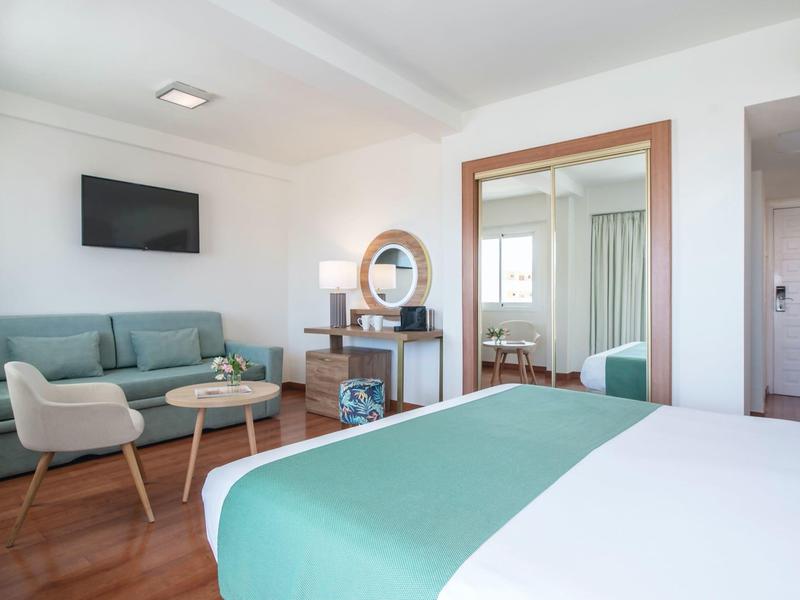 Helles Hotelzimmer mit Doppelbett, grünem Sofa, Sessel, TV und Holzmöbeln auf Holzboden.