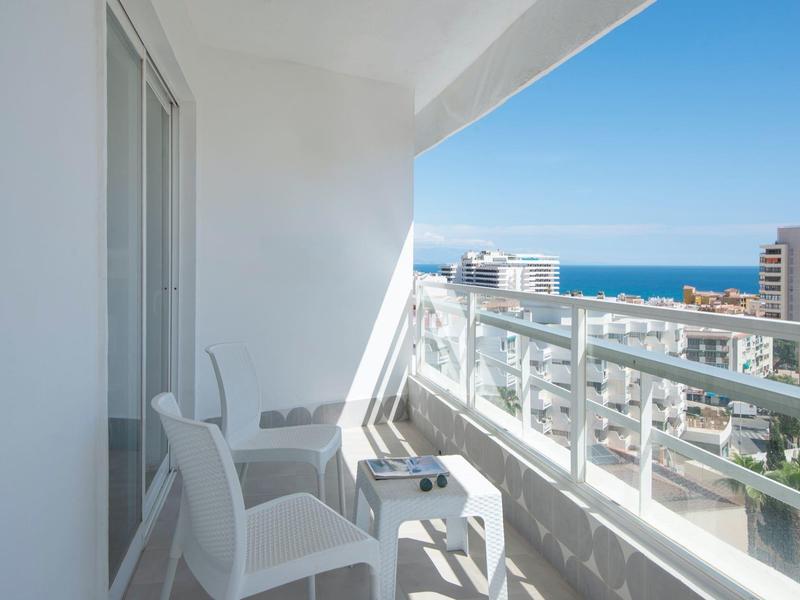 Balkon mit weißen Stühlen und Tisch, Glasgeländer, Blick auf Meer und Stadt an sonnigem Tag.