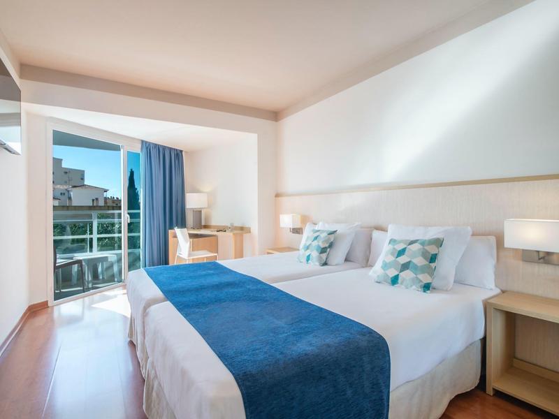 Helles Doppelzimmer mit weißen Wänden, blauer Bettdecke, Balkon und Fernsehgerät an der Wand.