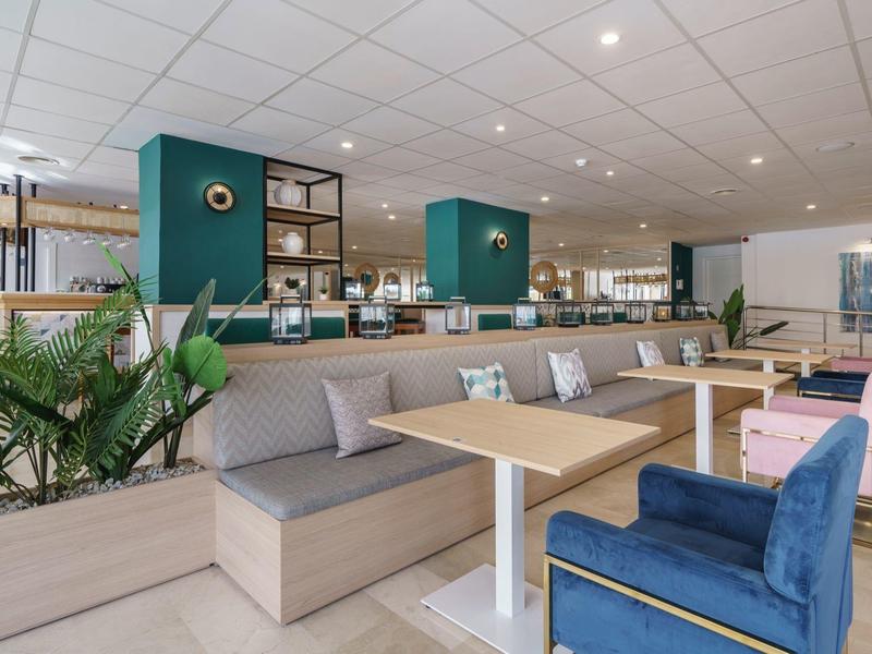 Moderner Cafébereich mit Holztheke, blauen Sesseln, grauen Sitzbänken und grünen Pflanzen.