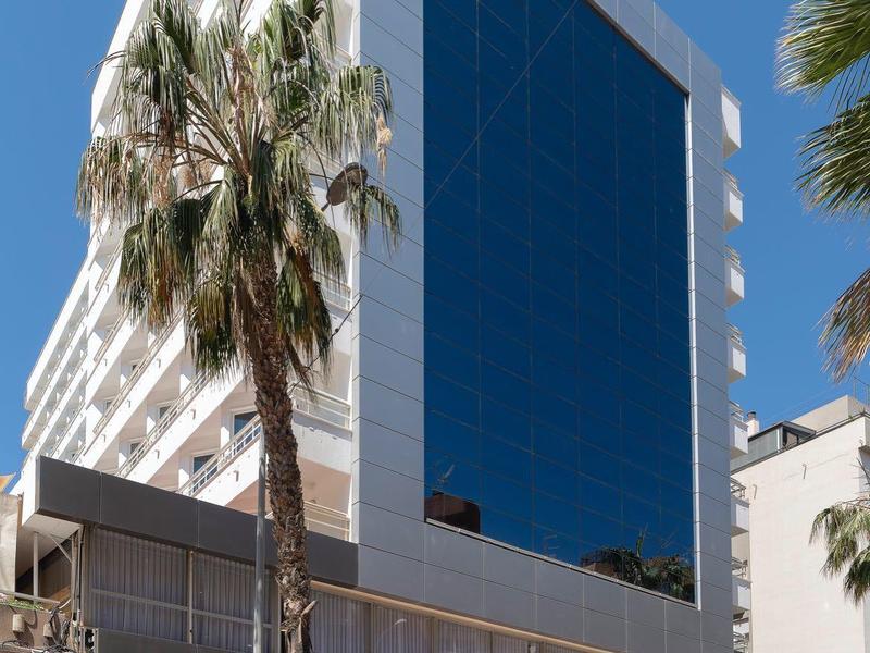 Modernes, weißes Gebäude mit großem, dunklem Glasfenster und Palmen vor klarem Himmel.