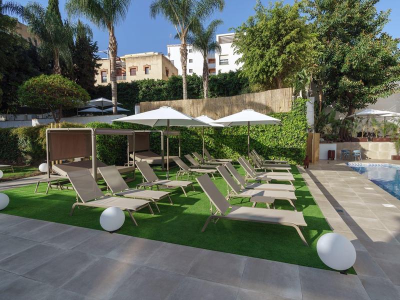 Poolbereich mit Liegestühlen, Sonnenschirmen und grüner Rasenfläche bei klarem Himmel.