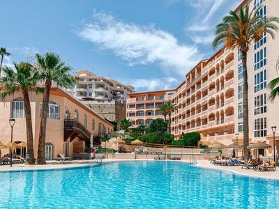 Hotel met groot buitenzwembad, palmbomen en meerlaagse gebouwen onder zonnige hemel.