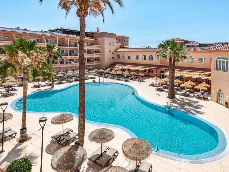 Groot zwembad met zonneparasols en ligstoelen bij een hotelresort met palmbomen.