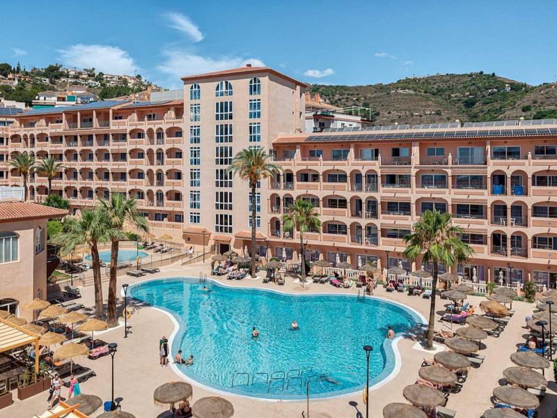 Groot hotelzwembad met ligstoelen, palmbomen en een meer verdiepend gebouw op de achtergrond.