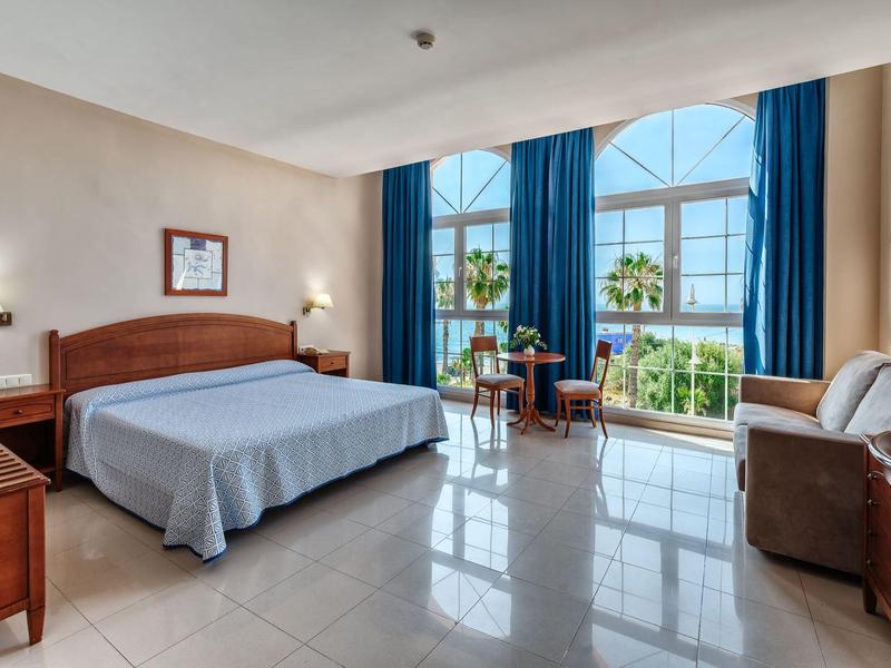 Licht hotelkamer met groot bed, bank en uitzicht op tropische planten.