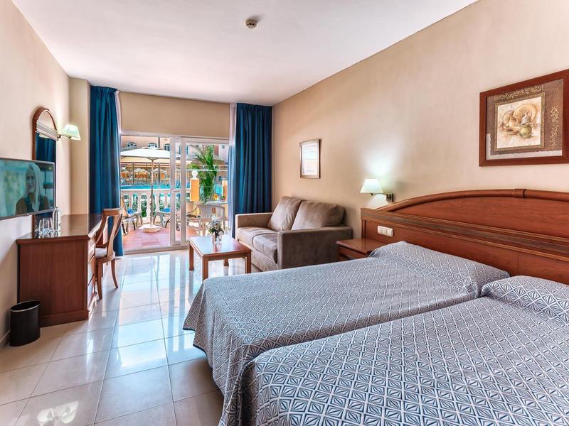 Lichte hotelkamer met twee eenpersoonsbedden, bank, bureau en balkon met uitzicht op het zwembad.